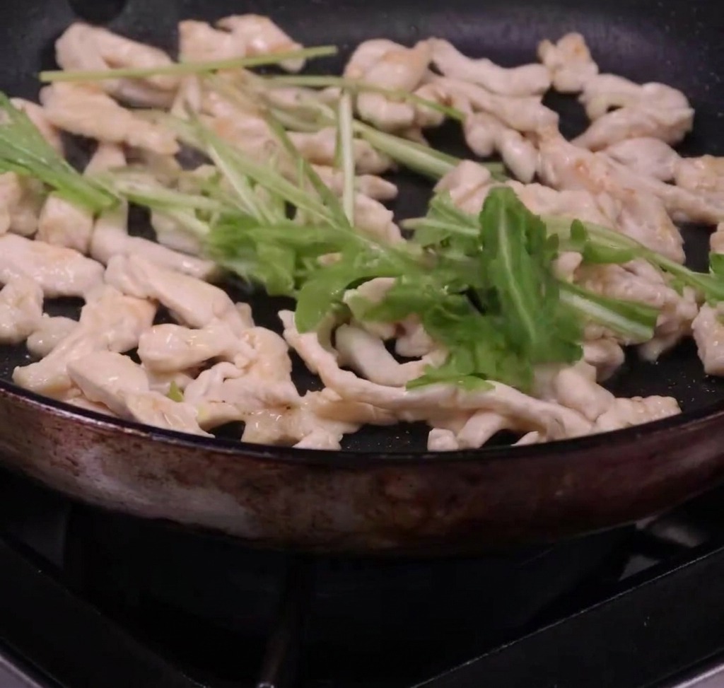Spaghetti and fresh arugula being tossed with cooked chicken strips in a pan.