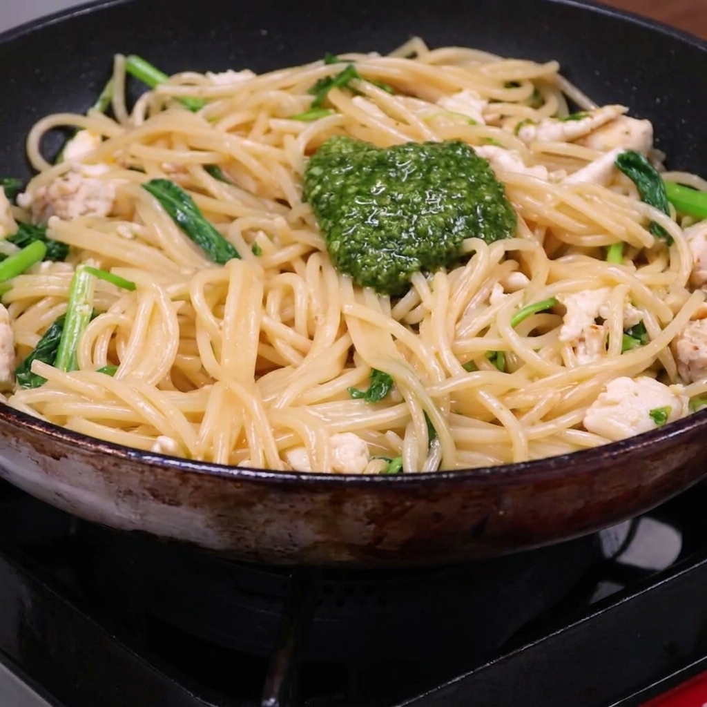 A dollop of vibrant green pesto sitting on top of a mixture of cooked spaghetti, chicken pieces, and arugula in a skillet.