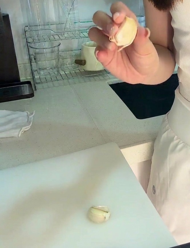 A hand holding a peeled clove of garlic over a white cutting board with a small amount of minced garlic visible.