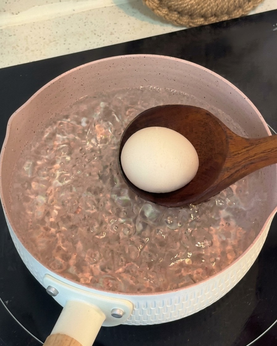 A wooden ladle gently lowering a whole egg into a pink pot of rapidly boiling water.