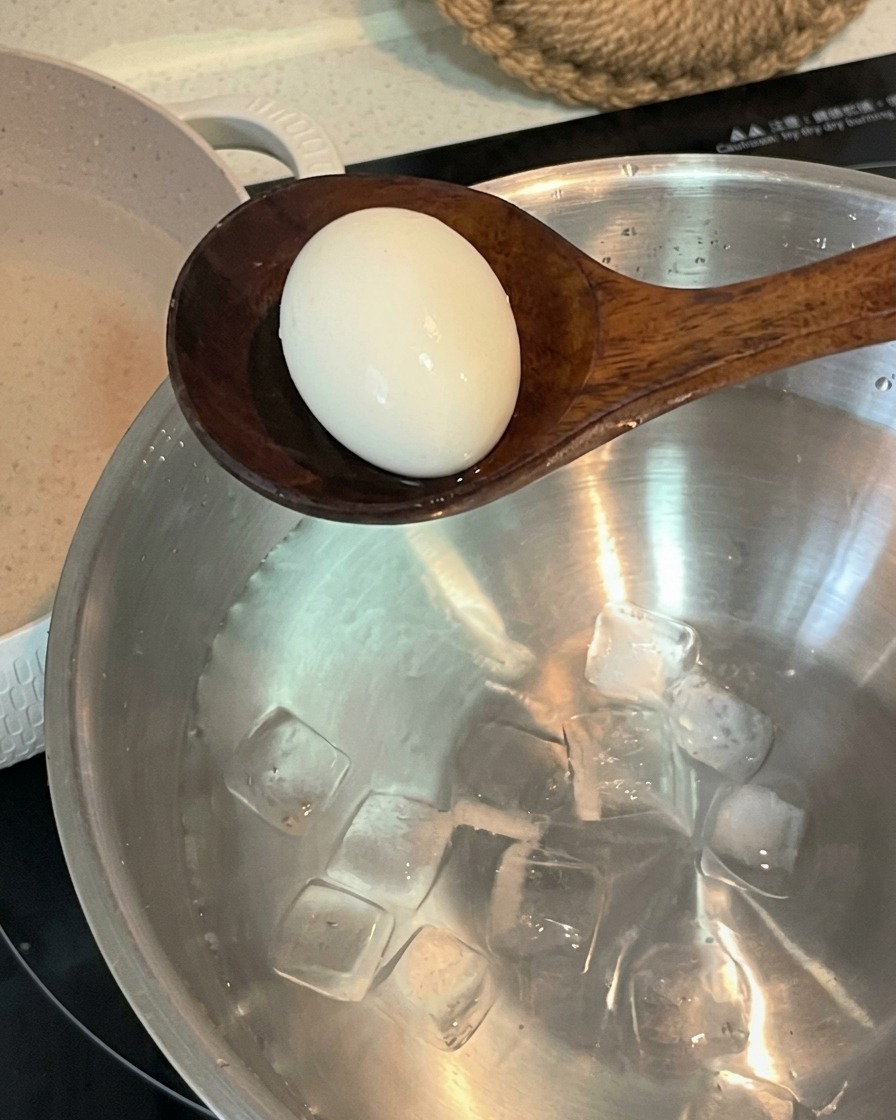 A wooden spoon transferring a boiled egg into a stainless steel bowl filled with cold water and ice cubes.