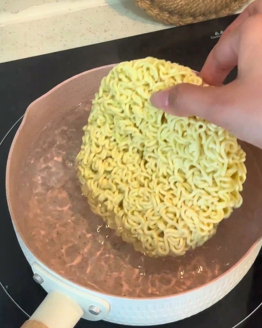 A hand placing a dry, round block of instant ramen noodles into a pot of boiling water.