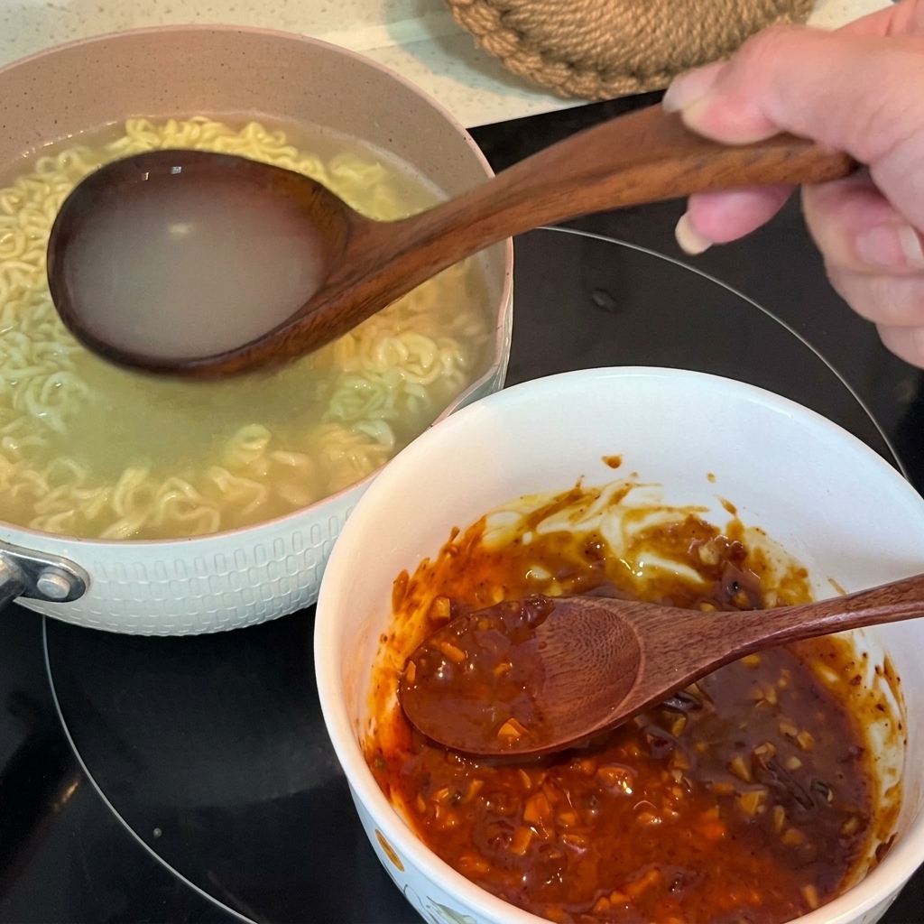 Hot noodle water being poured from a wooden ladle into a bowl containing a mixed seasoning paste.