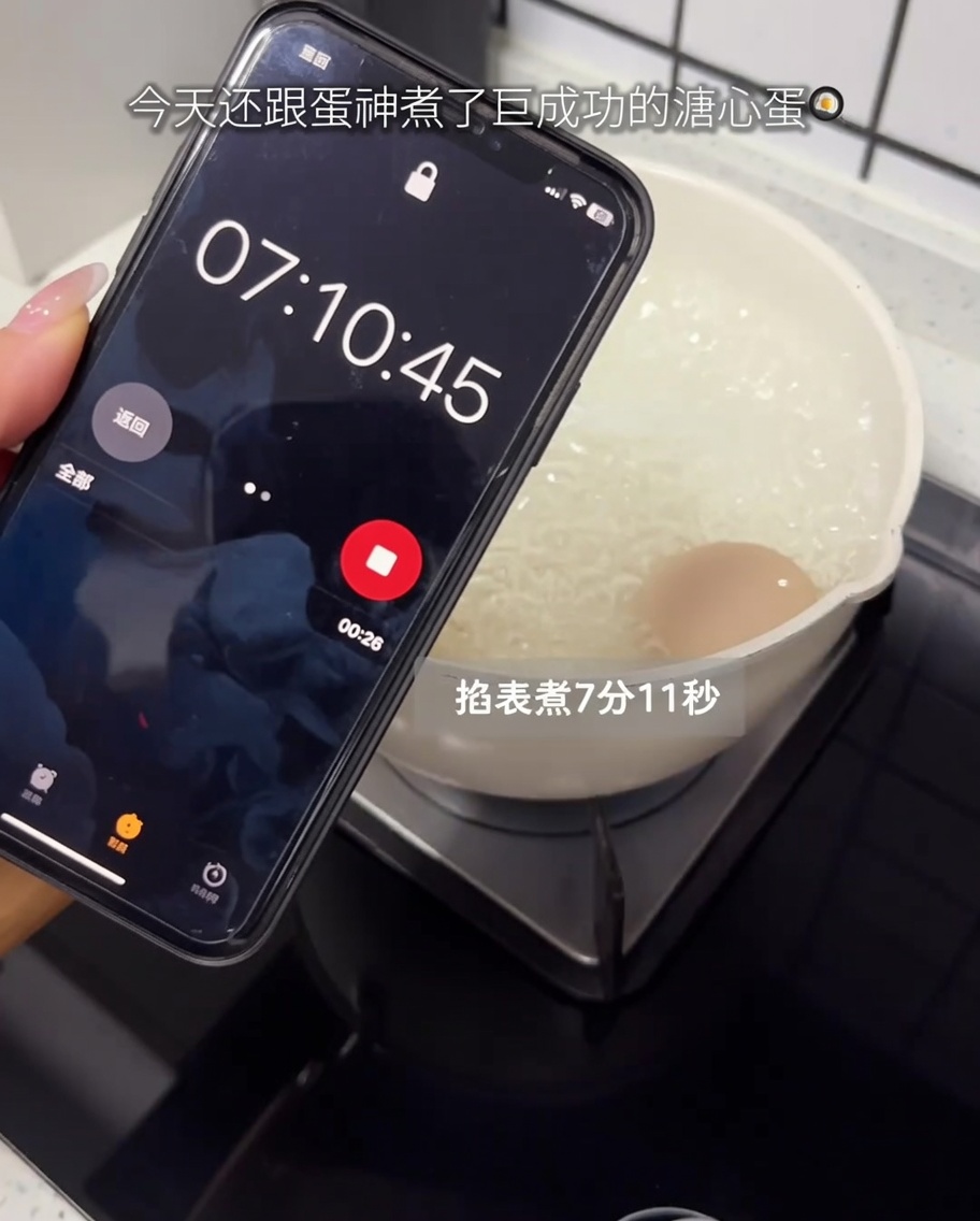 A raw egg boiling in a pot on a stovetop with a smartphone timer displayed in the foreground.