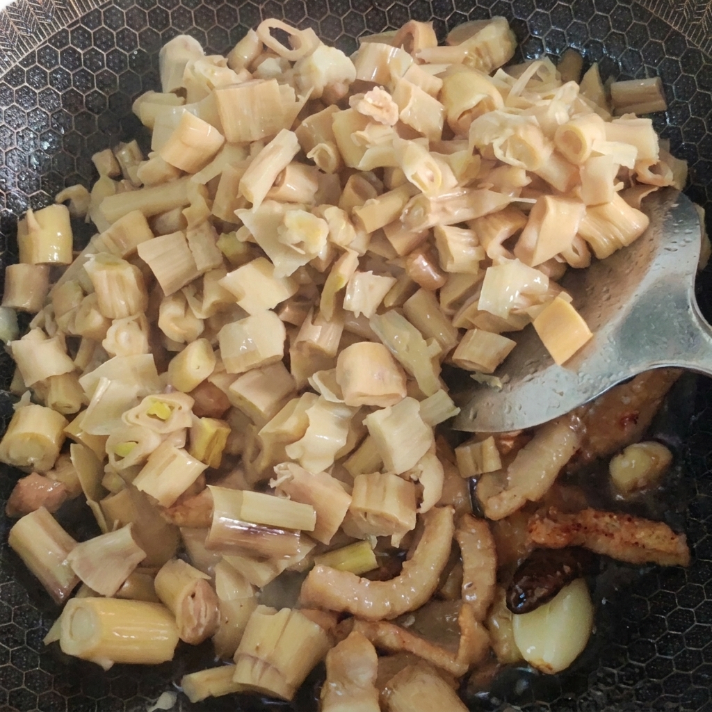 Water and pickled mustard greens being added to a wok filled with stir-fried pork and bamboo shoots.