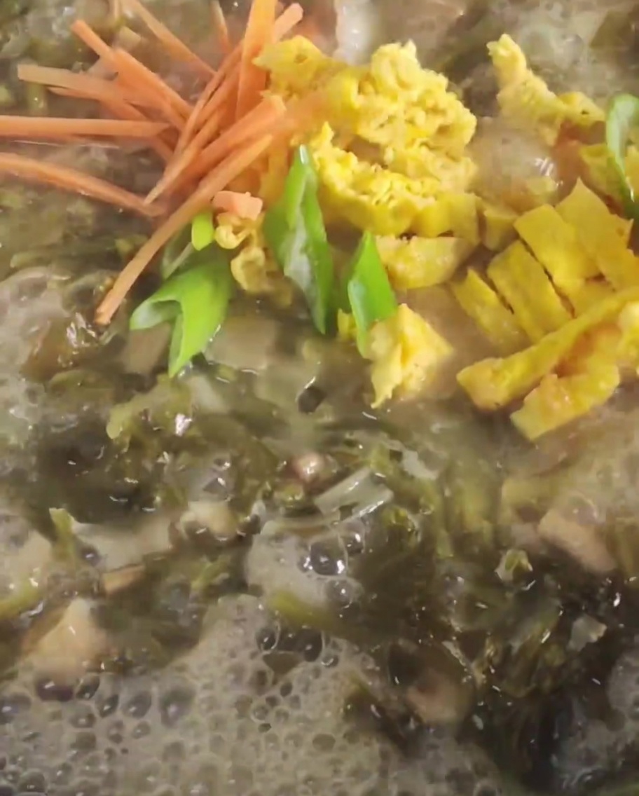 Carrots and sliced fried egg being added into a bubbling pot of broth with mustard greens.