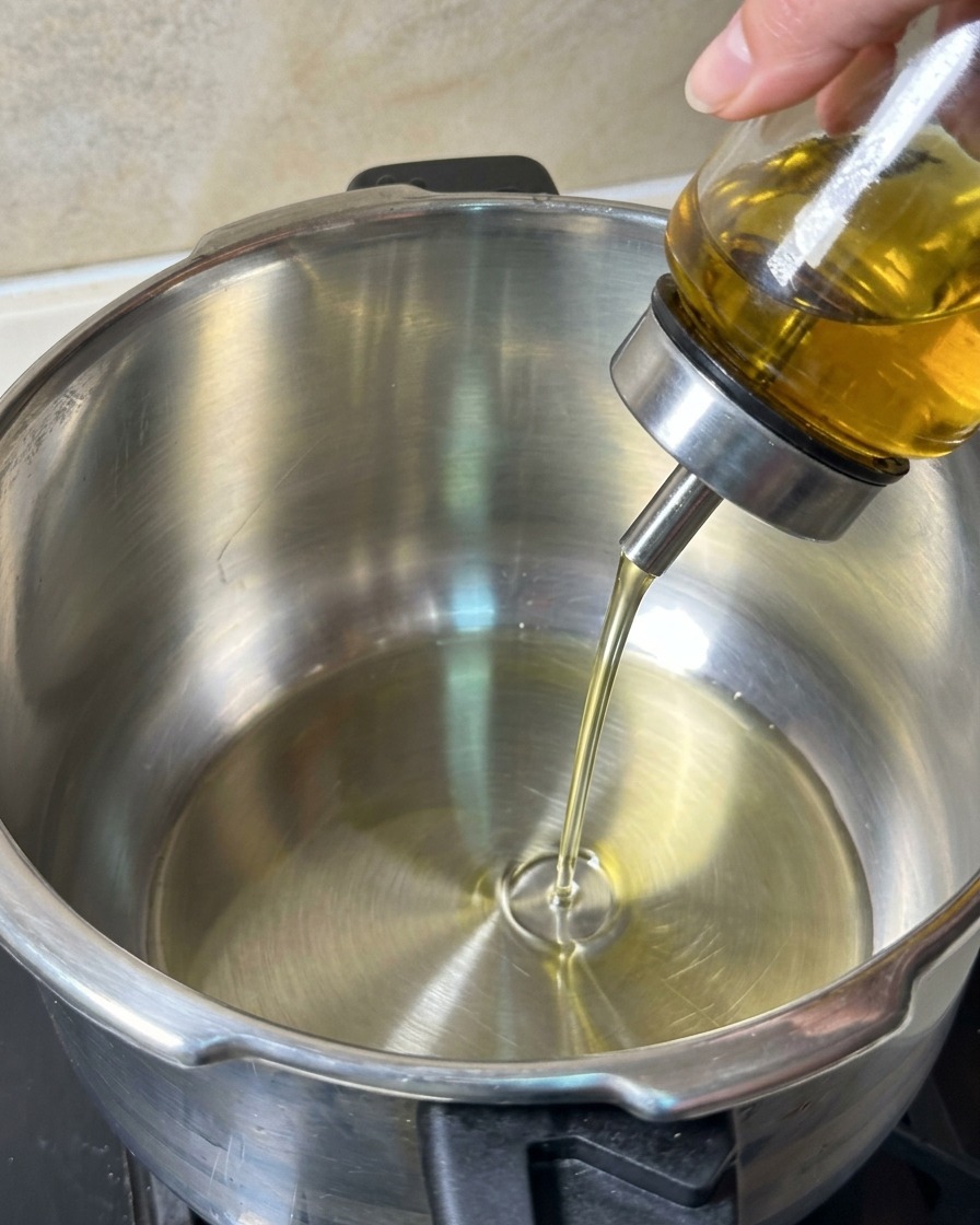 Cooking oil being poured from a glass dispenser into a clean stainless steel pot.