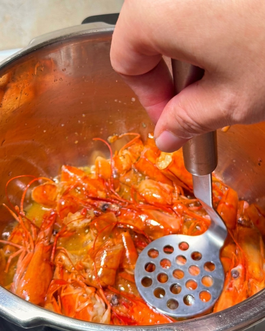 A potato masher pressing down on frying shrimp heads to extract golden orange oil.