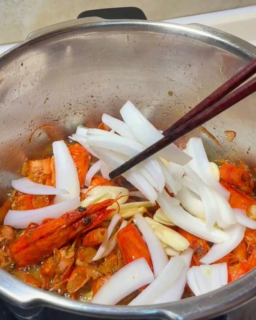 A close-up of garlic slices and onion strips being stirred into the fried shrimp shells in a metal pot.