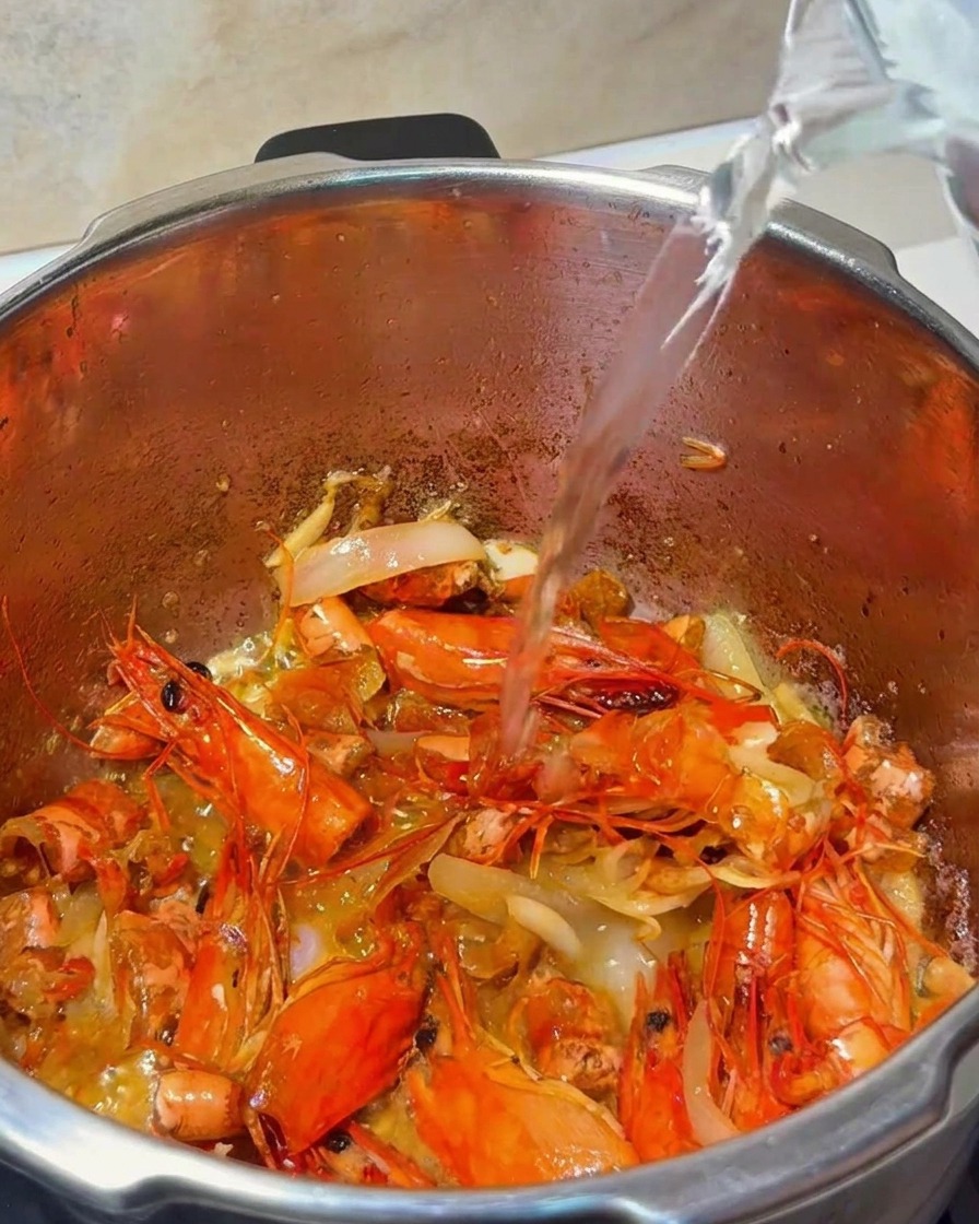 Water being poured from a glass pitcher into a pot containing shrimp shells and aromatics.