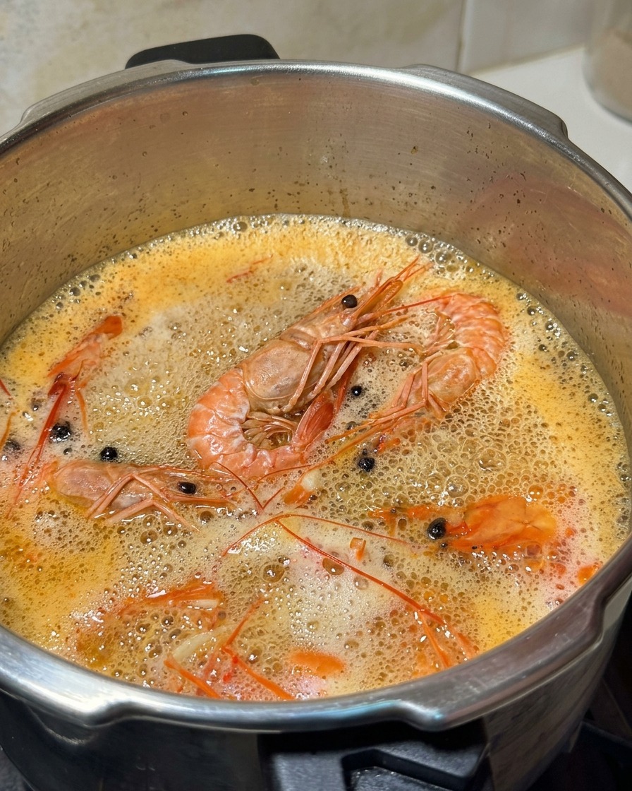 A metal ladle removing white foam from the surface of a simmering orange shrimp broth.