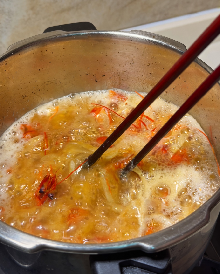 A knob of butter melting into a pot of simmering orange shrimp stock, being stirred with chopsticks.