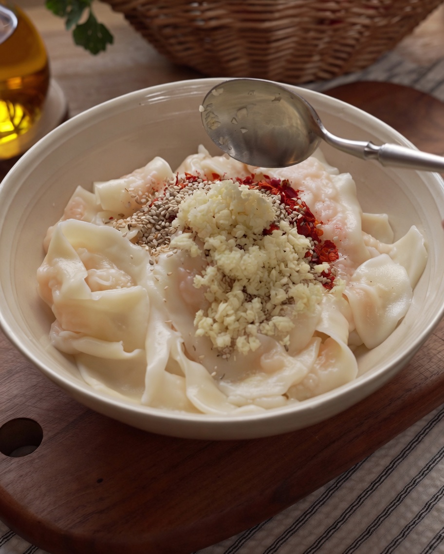 Hot oil being poured from a silver ladle over the spices and minced garlic on top of the cooked noodles, causing them to sizzle.