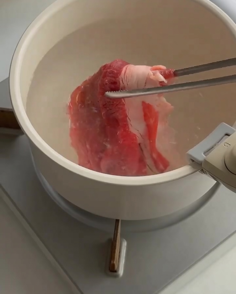 Thin slices of raw beef being lowered into a pot of boiling water using a pair of tongs.