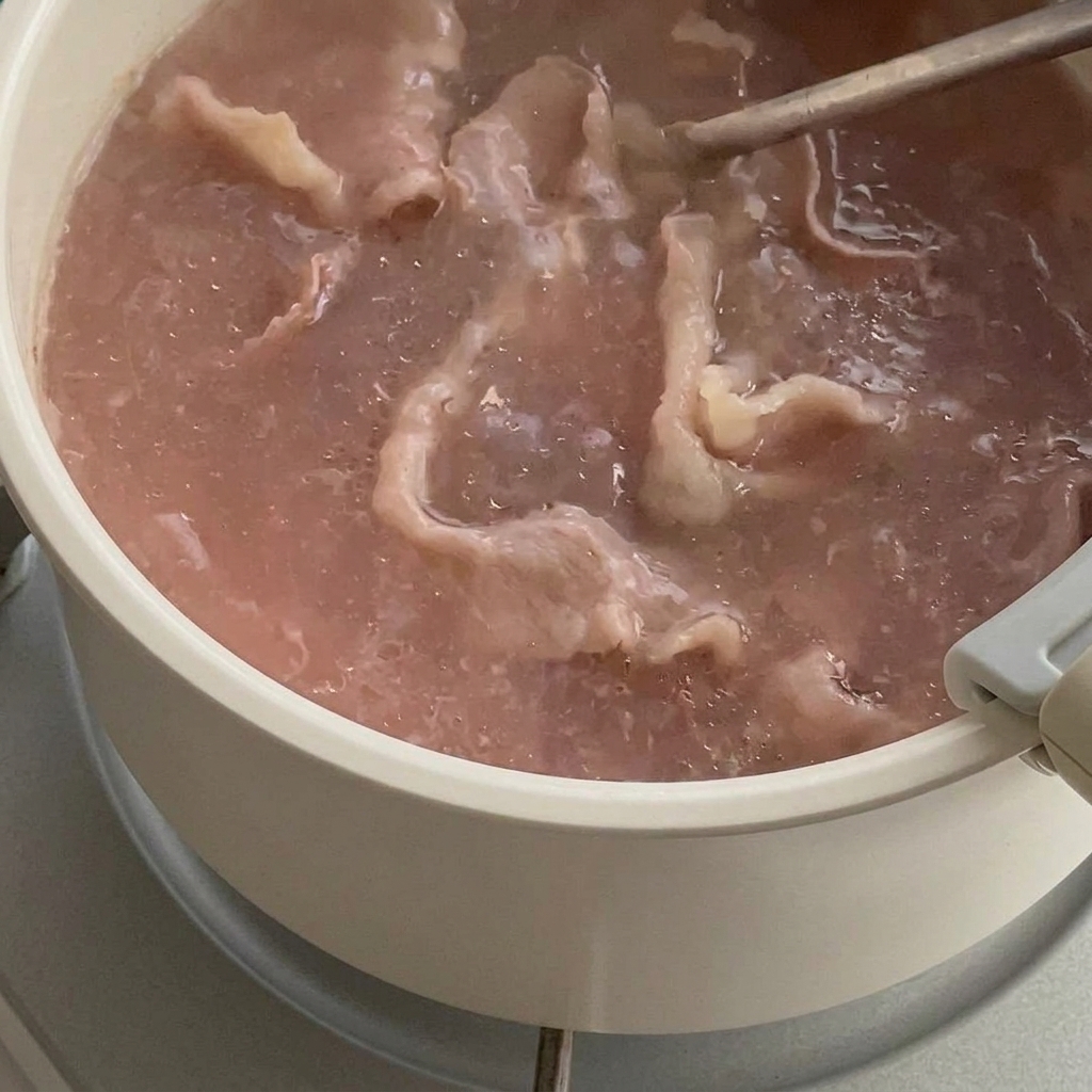Partially cooked thin beef slices boiling in a pot of water, being lifted with a wire strainer.