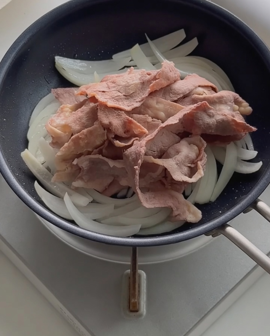 Blanched beef slices resting on top of sautéed onions in a black skillet.