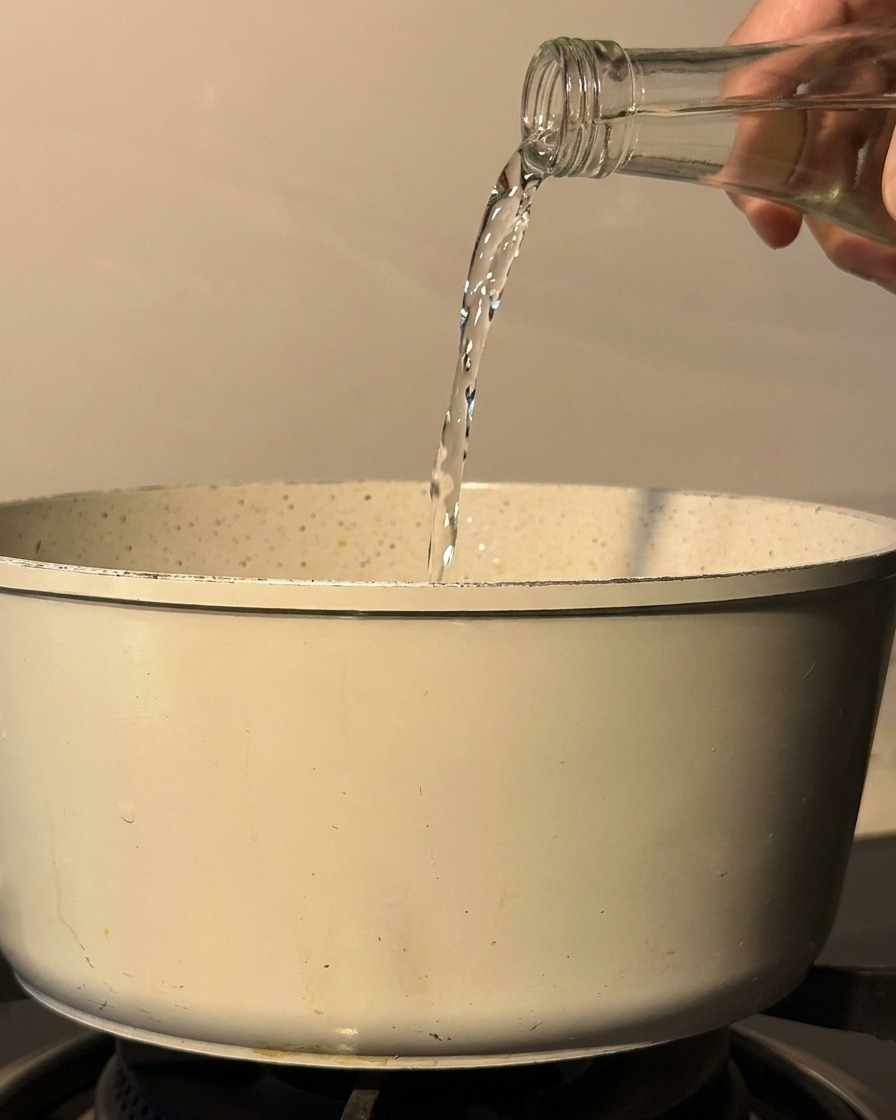 Sake and mirin being poured from a glass bottle into a white cooking pot on a stovetop.