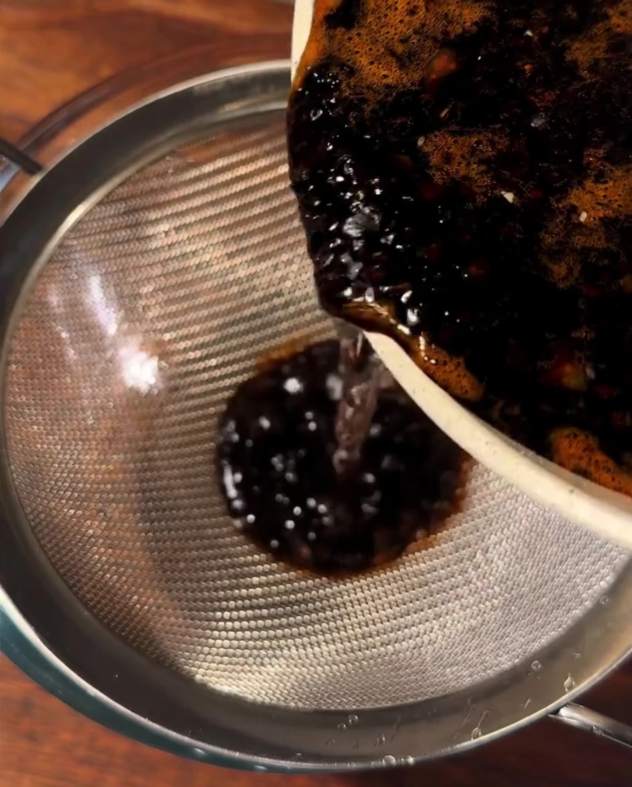 Dark brown sauce being poured through a metal sieve into a glass bowl.