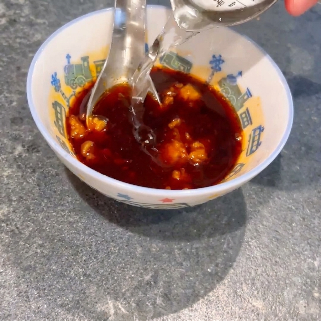 Water being poured into a small bowl containing dark soy sauce and miso lumps, with a spoon stirring the mixture.