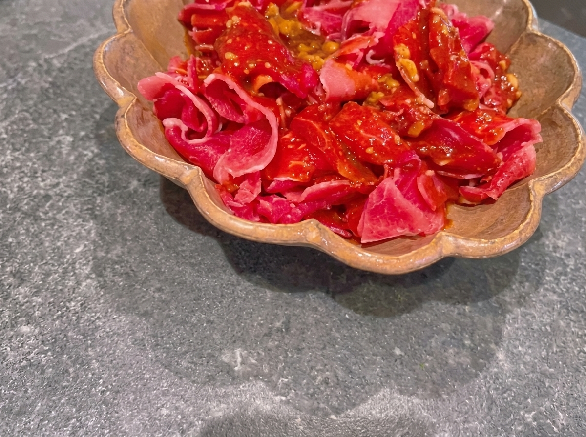 Thinly sliced raw red beef coated in a dark miso marinade inside a brown scalloped dish.