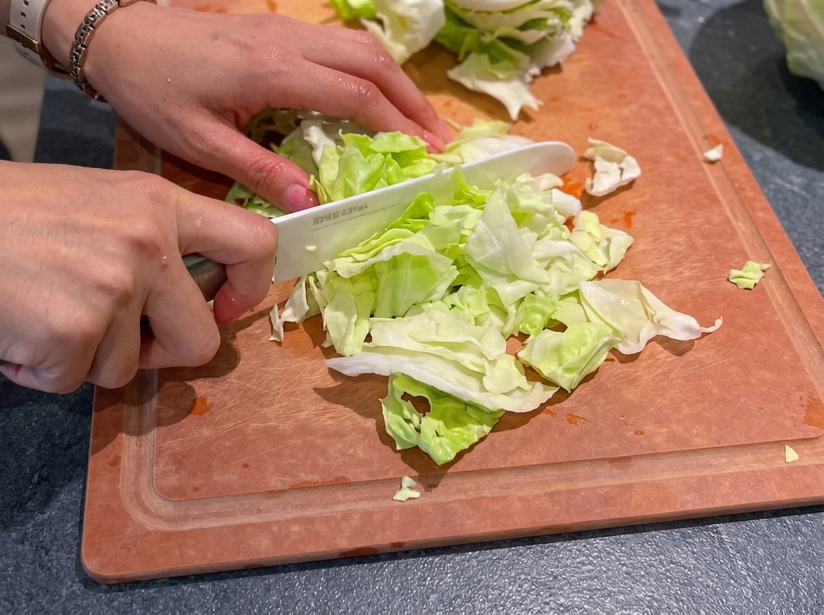 Hands using a white ceramic knife to chop fresh green cabbage into small pieces on a wooden cutting board.
