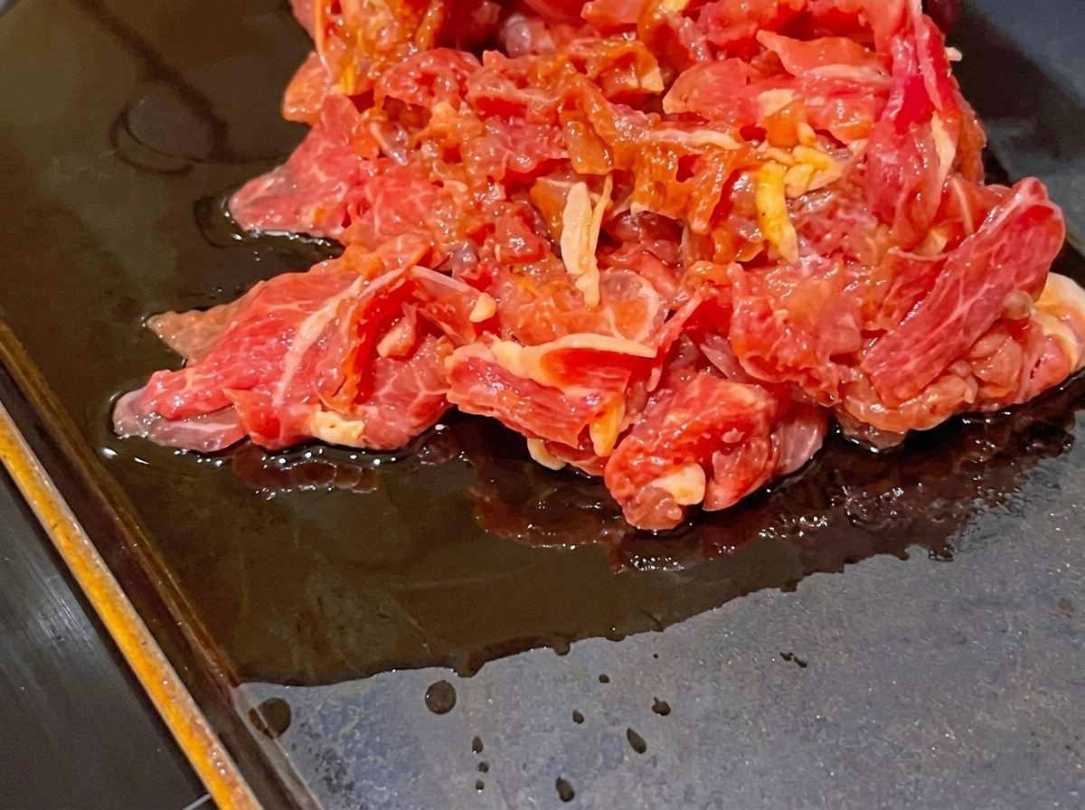 A pile of marinated raw red beef slices sitting on a hot, dark, flat cooking pan.