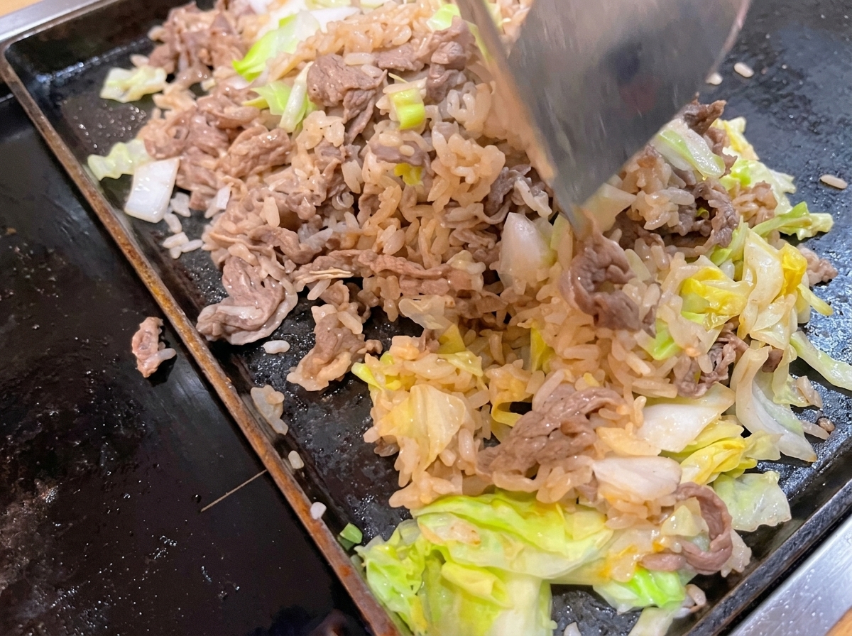 A metal spatula tossing and mixing cooked beef, white rice, and green cabbage together on a hot flat pan.