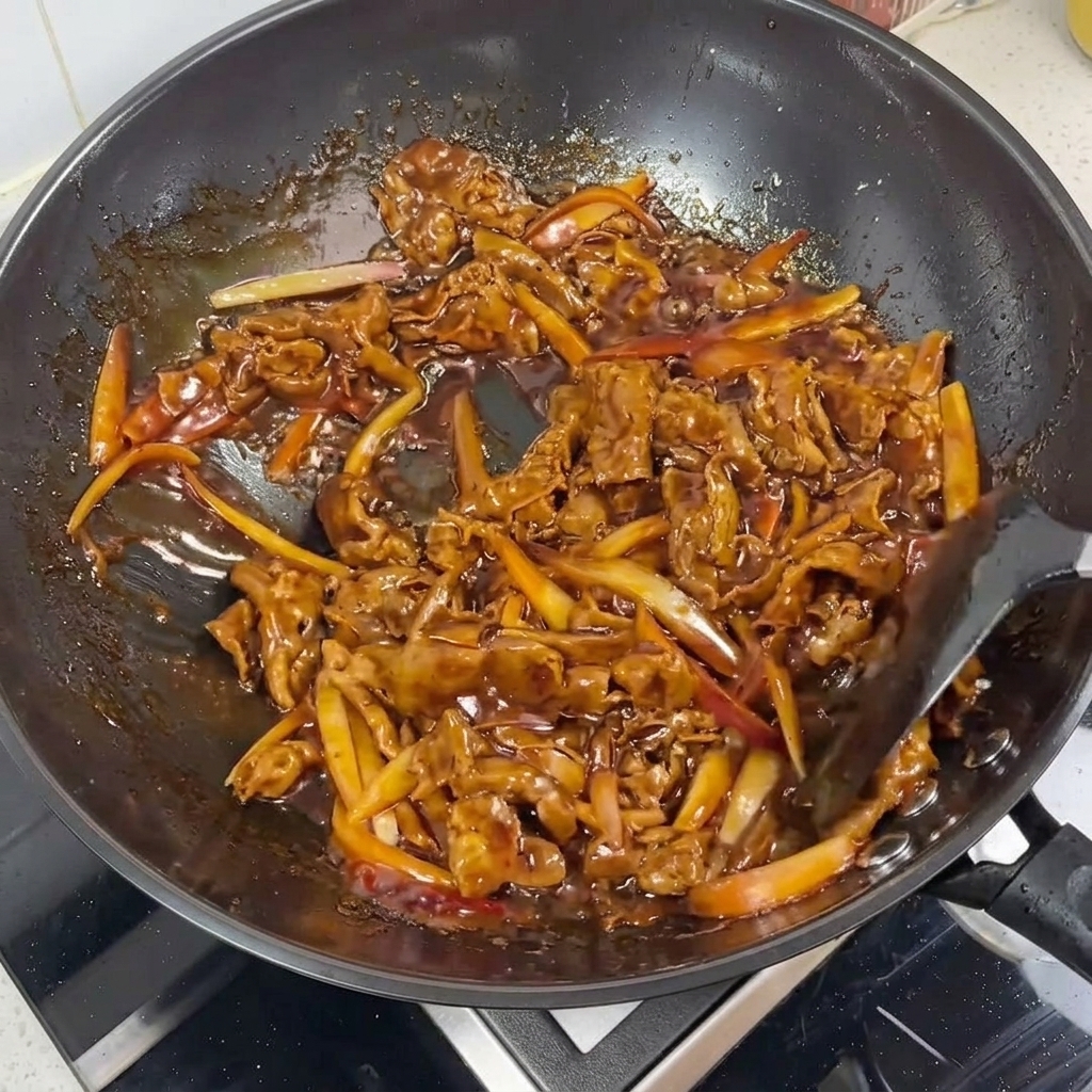 A close-up of beef slices and red onion strips being glazed in a dark, glossy teriyaki sauce in a wok.