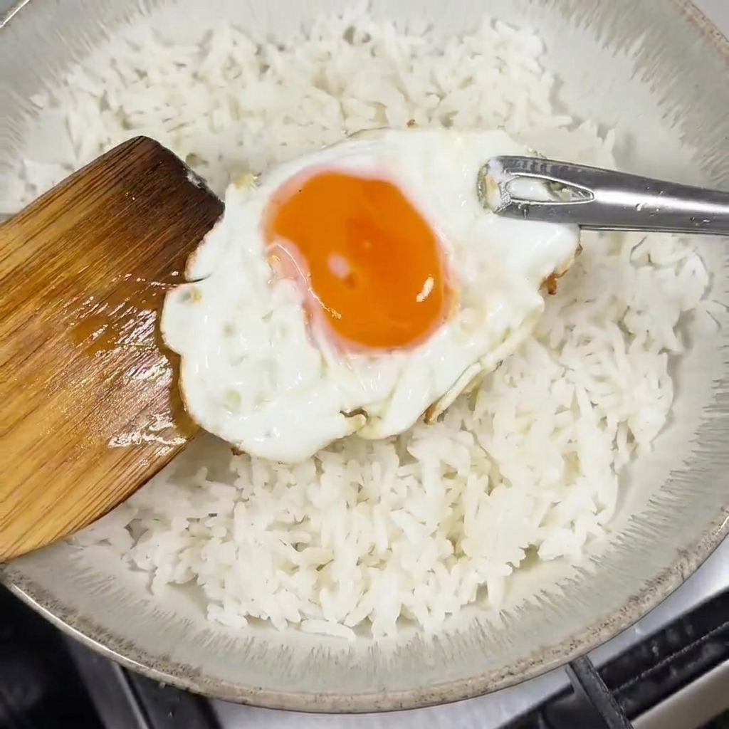 A wooden spatula placing a fried egg with a runny yolk onto a bowl filled with white rice.
