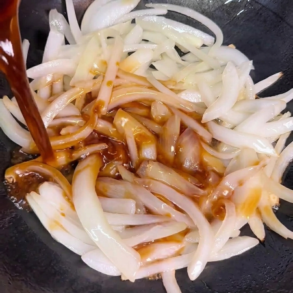 Sauce being poured over the sautéed onions in a wok.