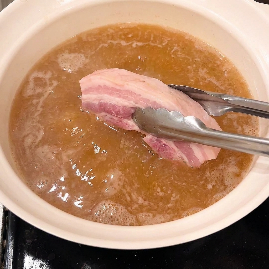 Tongs placing blanched pork belly pieces into a pot of simmering brown braising liquid.
