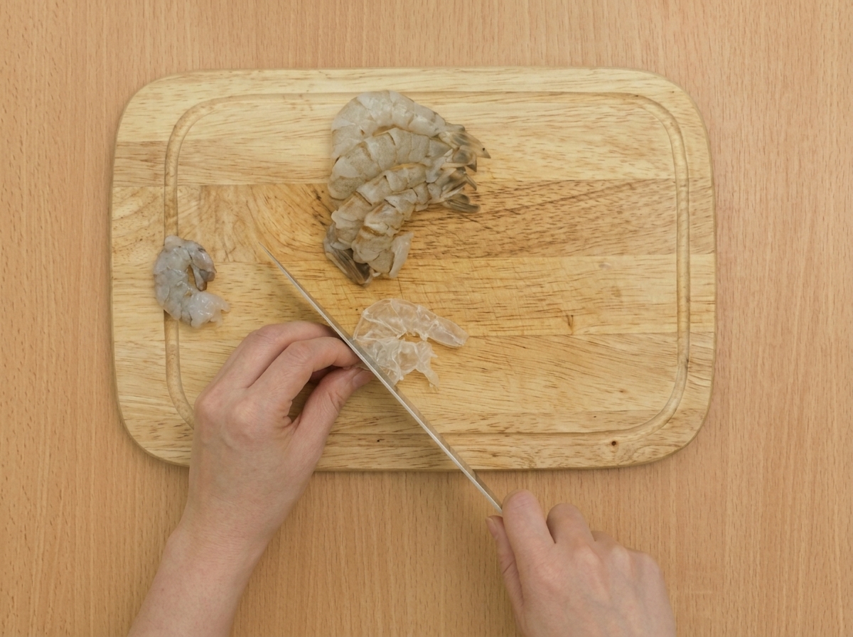 A close-up of a knife making shallow cuts along the belly of a raw shrimp on a wooden cutting board.