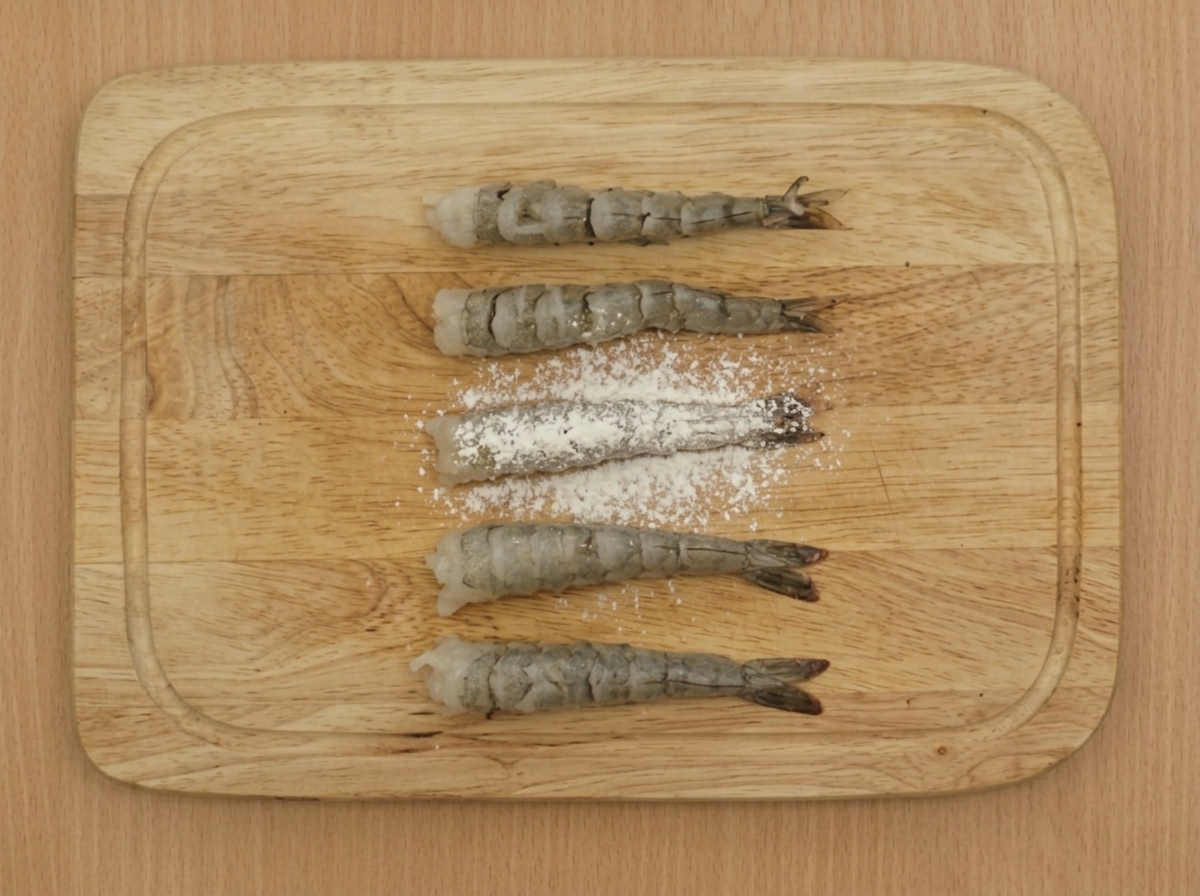 A sifter dusting low-gluten flour over several prepared shrimp arranged on a wooden board.