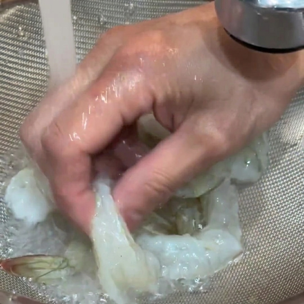 Raw shrimp being rinsed under running tap water in a colander.