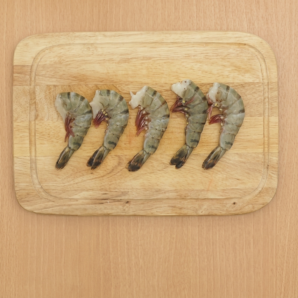 Five cleaned shrimp on a wooden cutting board with their tails still attached.