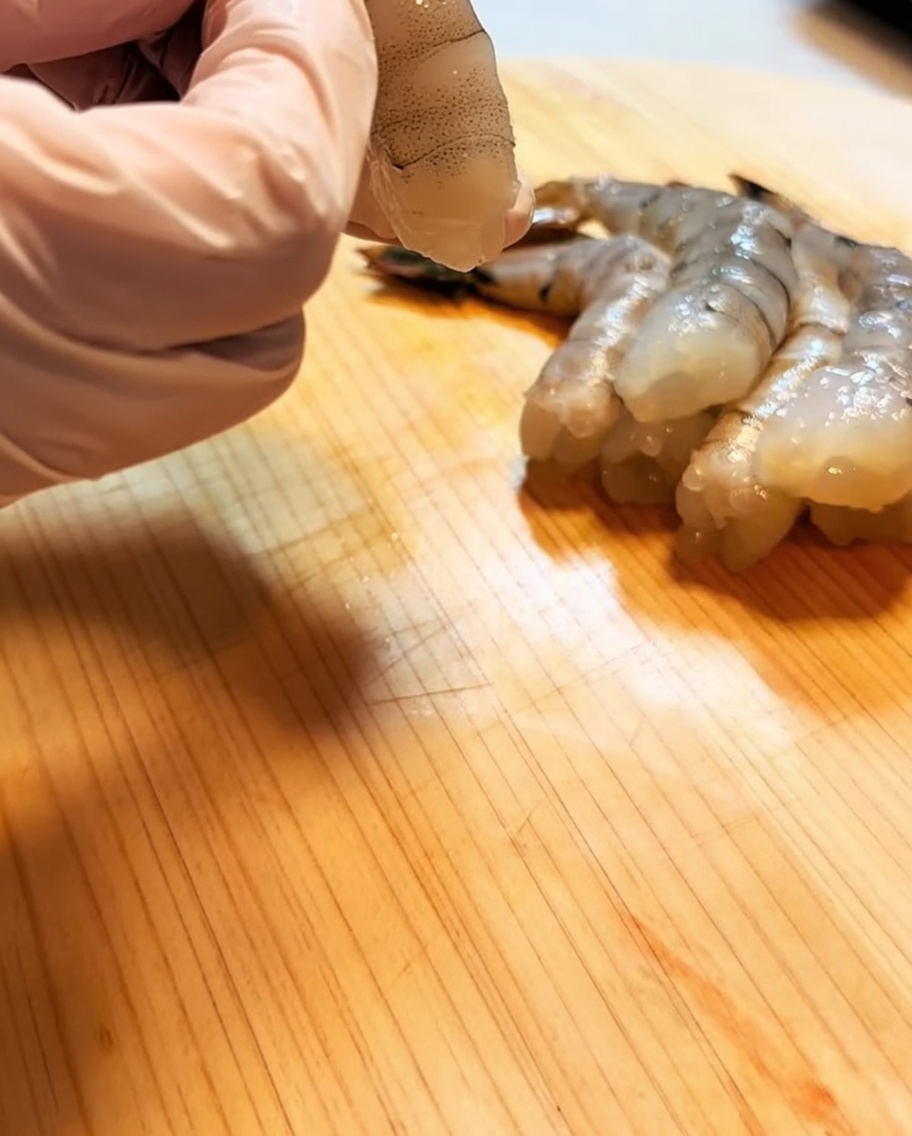 A hand wearing a pink glove holding a cleaned, deveined, and prepped shrimp on a wooden surface.