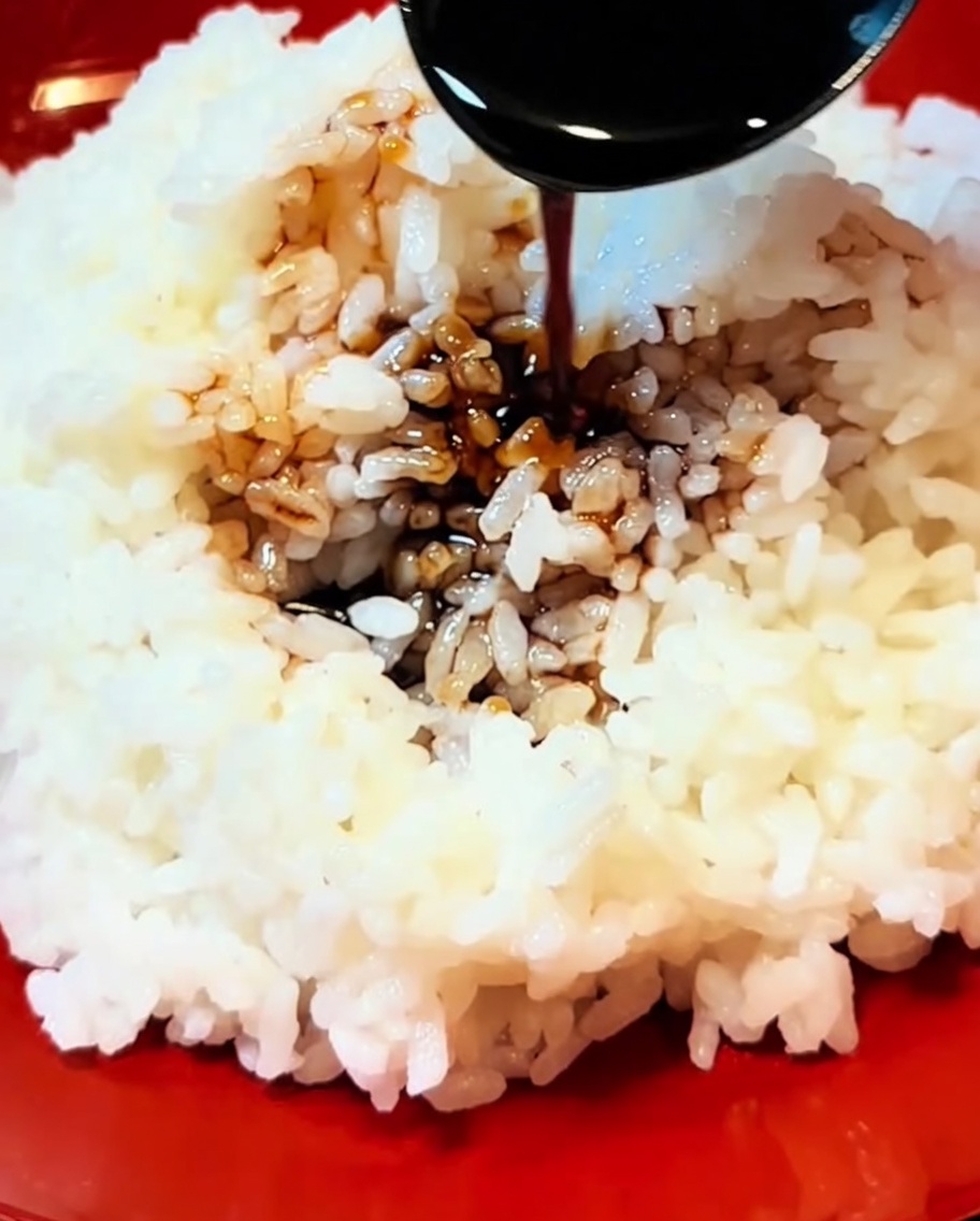 A close-up view of dark, thickened sauce being poured from a spoon onto a serving of fluffy white rice in a red bowl.