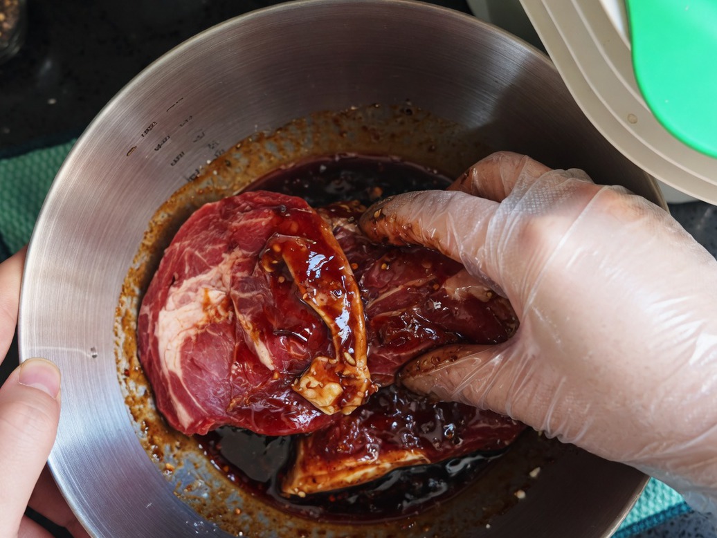 Hand in a plastic glove mixing raw pork chops with marinade in a metal bowl.