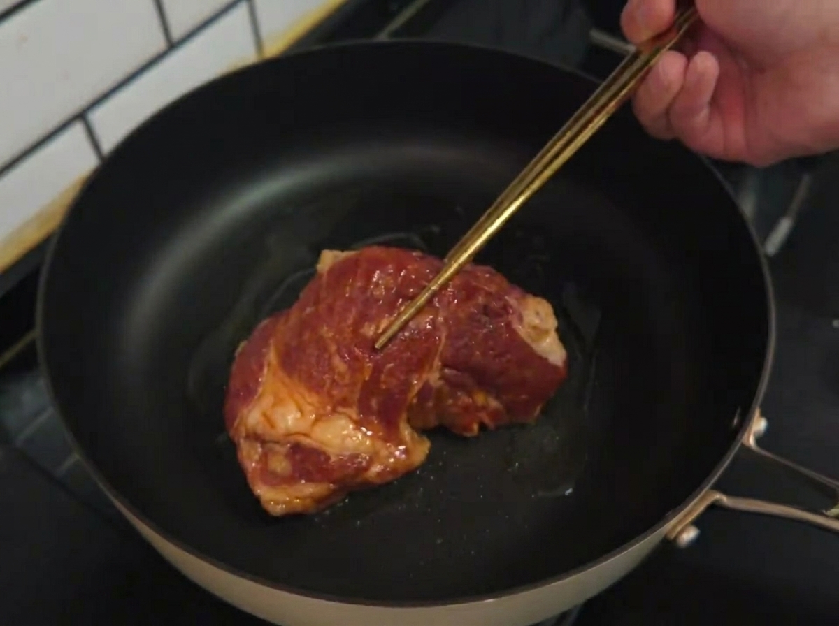 Raw marinated pork chop being laid into a hot pan with cooking oil.