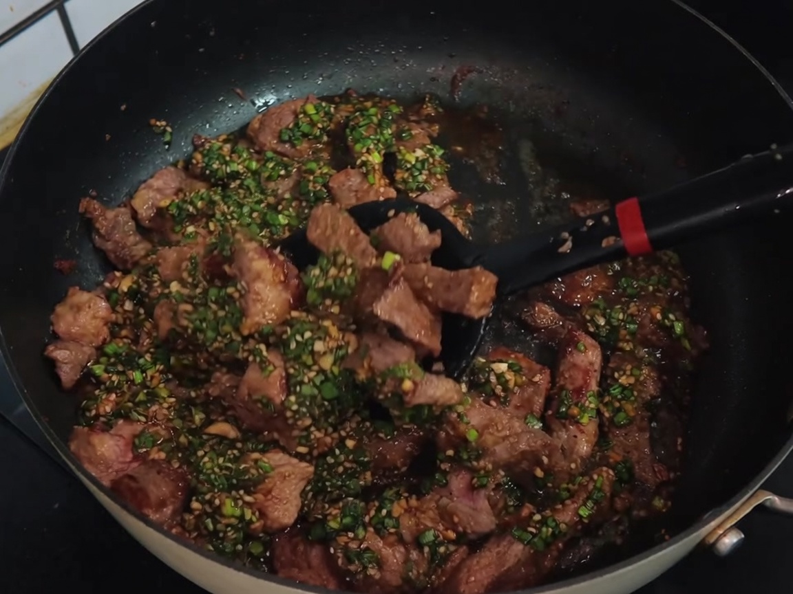 A black spatula mixing sliced pork chops with a green scallion sauce in a non-stick pan.