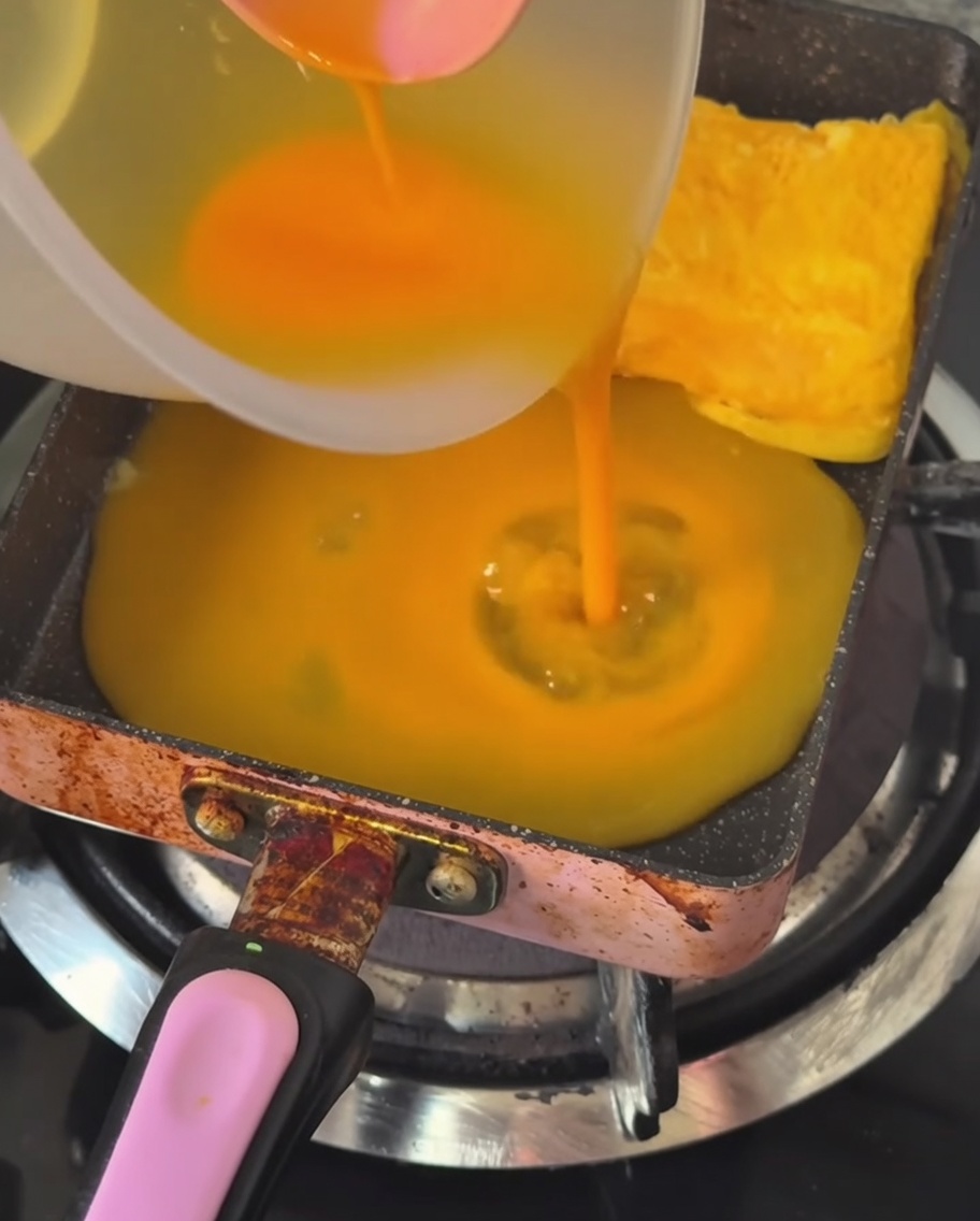 A thin layer of whisked egg being poured into a hot, greased rectangular pan.