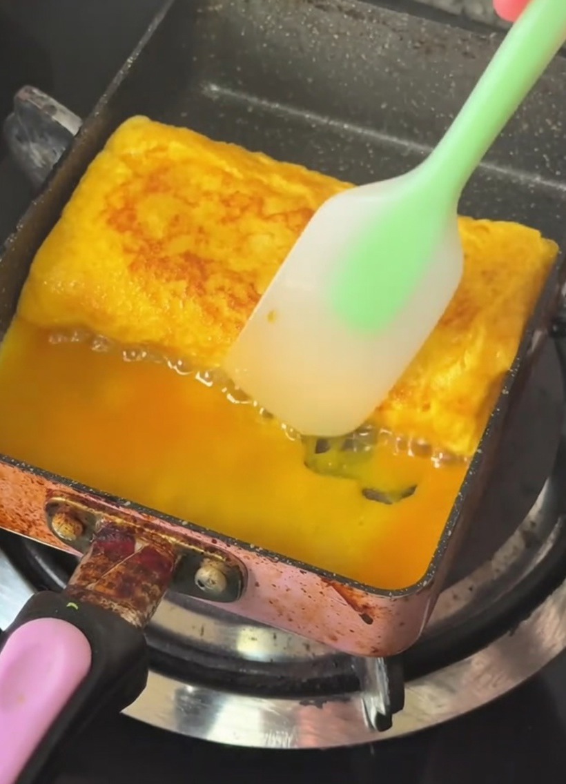 A rectangular tamagoyaki pan with a layered, golden egg roll being folded using a silicone spatula.
