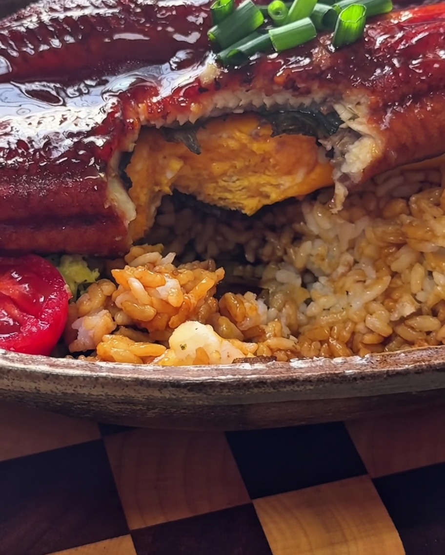 A bright yellow, folded tamagoyaki omelet being placed onto a base of seasoned rice in a bowl.