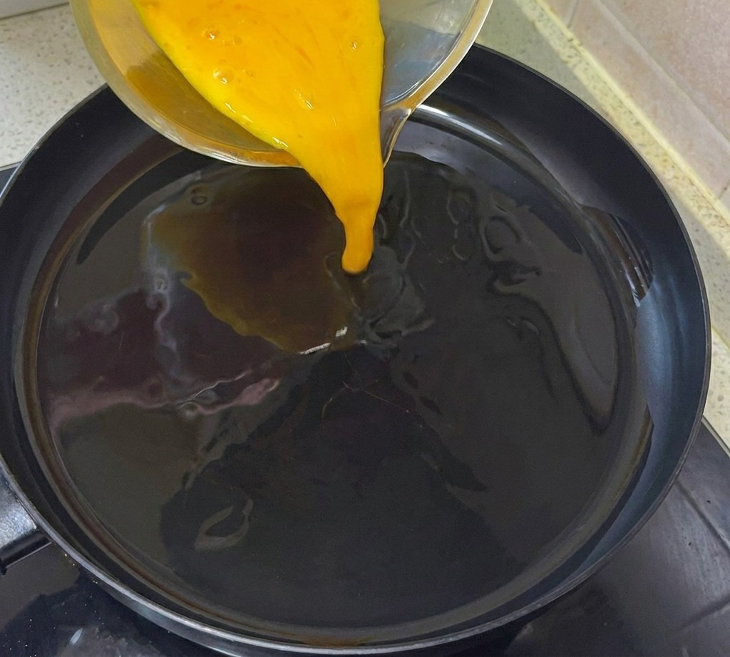 A stream of beaten egg mixture being poured into a preheated, oiled frying pan.