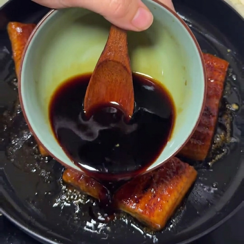 A dark, savory sauce being poured from a small bowl over golden-brown grilled eel fillets sizzling in a pan.