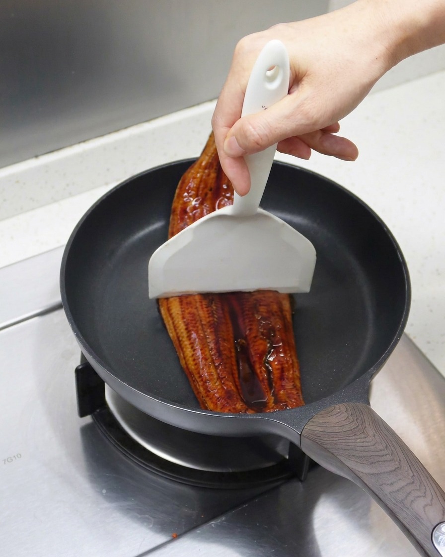 The grilled eel being divided into two pieces with a white spatula inside a frying pan.