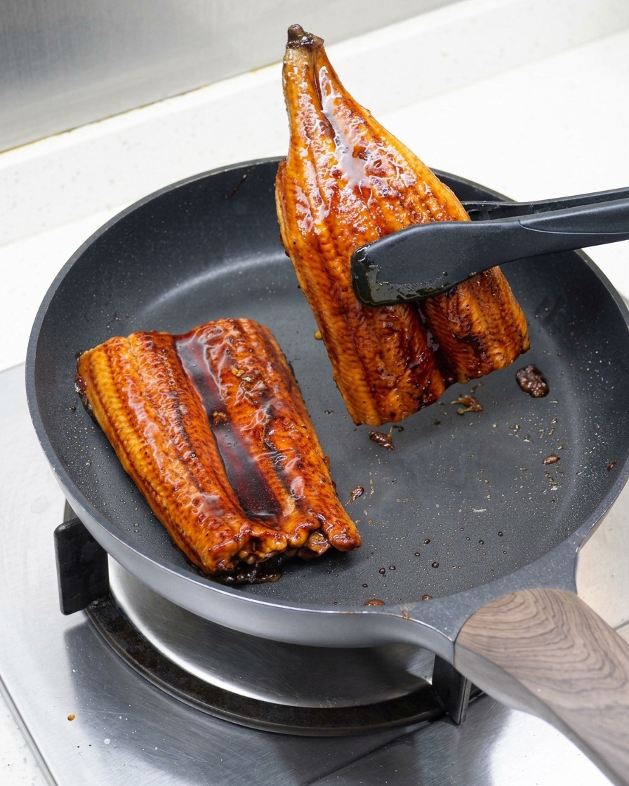 Two pieces of grilled eel being flipped in the pan to ensure even browning.