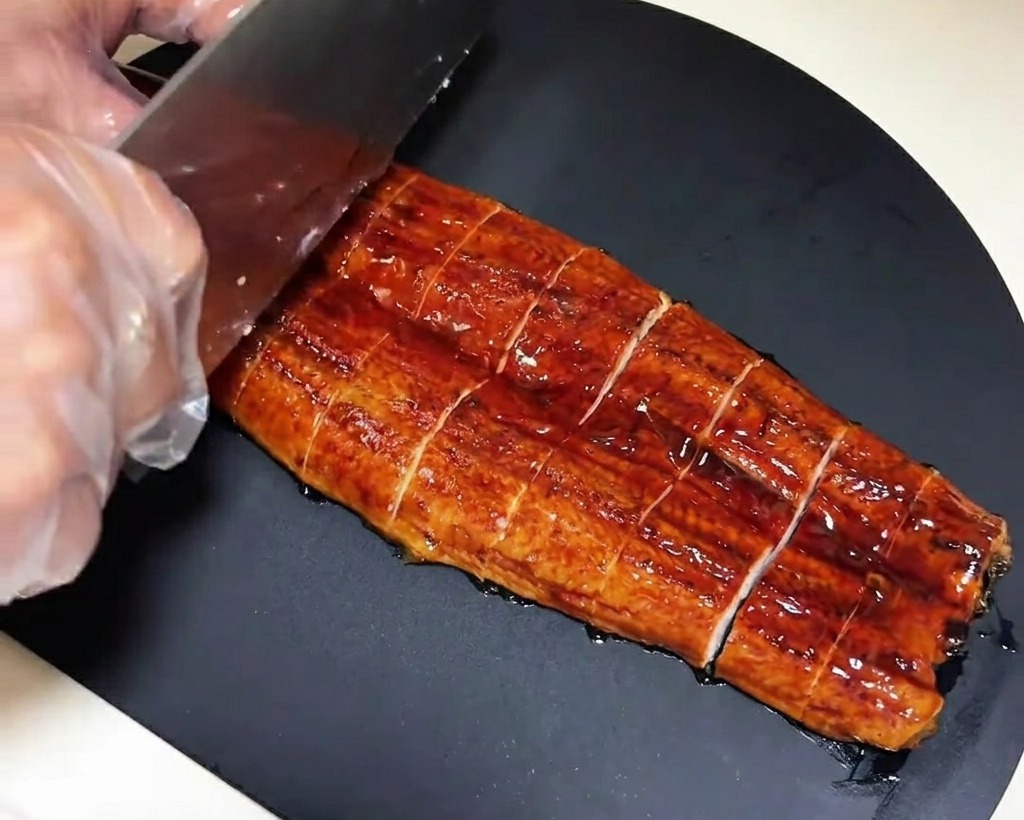 A pre-grilled eel fillet being cut into uniform, rectangular pieces on a dark cutting board.