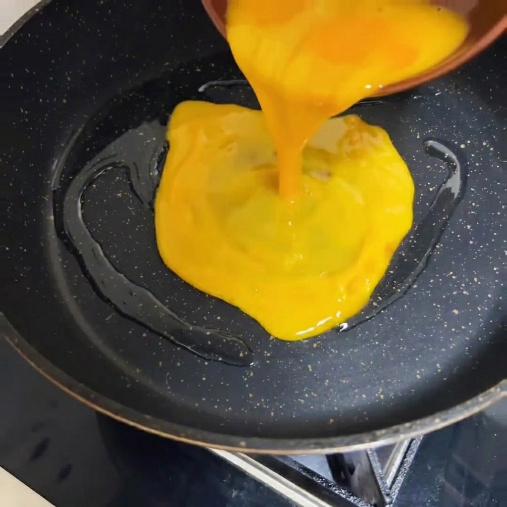 Beaten egg liquid being poured into a hot, dark-colored pan.