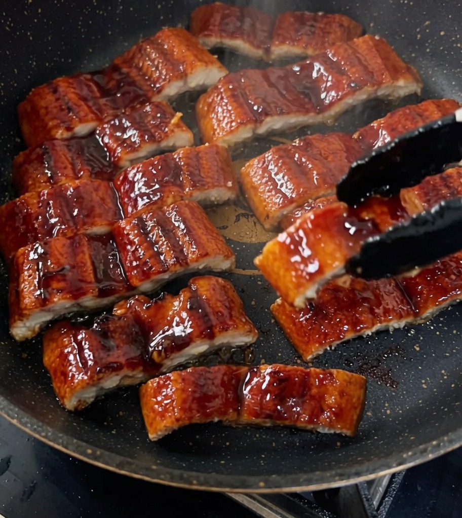 Slices of grilled eel being glazed in a pan with a dark, rich sauce.